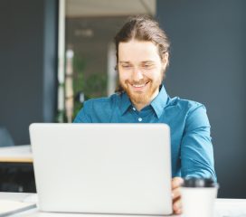 Happy young male office employee in shirt sitting at desk in front of laptop computer and looking at screen with smile, enjoying work in office. Cheerful man using digital technologies at workplace